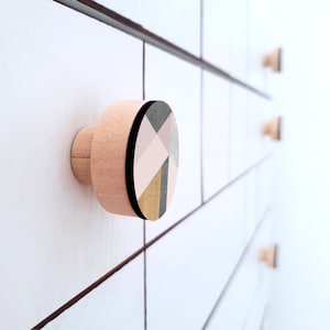 Puede incluir: Primer plano de un tirador de armario de madera con diseño geométrico. El tirador es de color rosa claro con un borde negro y una cara circular con un patrón de formas rosas, grises y doradas. Otros tiradores son visibles en el fondo.