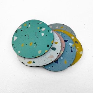 May include: A set of six colorful, round coasters with a terrazzo design. The coasters feature a variety of colors, including teal, gray, pink, blue, and yellow, with a black border. The coasters are arranged in a fan shape.