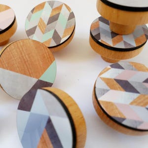 May include: Set of wooden drawer knobs with geometric patterns in shades of grey, pink, and mint green. The knobs have a black rim and are made of wood.