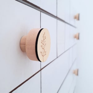 May include: A light pink wooden drawer knob with a black edge and a botanical design. The knob is mounted on a white surface with other knobs visible in the background. The design features a simple, elegant aesthetic.