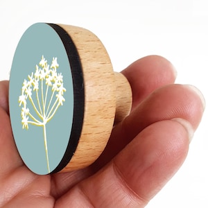 Peut inclure: Un bouton de meuble rond en bois avec une face bleu clair ornée d'un motif floral blanc et jaune. Le bouton a une bordure noire et une finition en bois naturel. Le bouton est tenu dans une main.