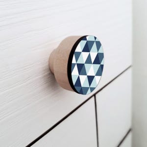 Op de afbeelding: Een houten kastknop met een geometrisch ontwerp. De knop heeft een natuurlijke houten basis en een cirkelvormig vlak met een patroon van blauwe en witte driehoeken. Een zwarte band omlijnt het vlak. De knop is op een witte kast gemonteerd.