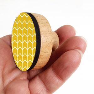 Op de afbeelding: Een ronde houten stempel met een geel en wit chevron-patroon. De stempel wordt in een hand gehouden.