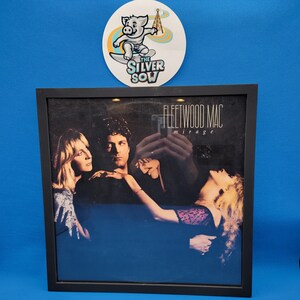 May include: Framed Fleetwood Mac "Mirage" album cover art. The album cover features three people in a dark setting with the album title in white text. A circular sticker with a pig surfing is above the frame.
