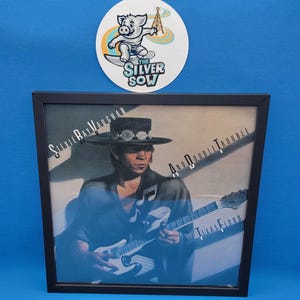 May include: Framed print of Stevie Ray Vaughan playing guitar, with the album title "Texas Flood" visible. The frame is black, and a sticker with a pig surfing is above the frame. The sticker reads "The Silver Sow".