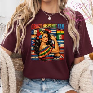 May include: A maroon t-shirt featuring a graphic of a woman flexing her arm, adorned with flowers and a rainbow-colored shawl. The text "CRAZY HISPANIC FAN" is above the graphic, with various flags in the background.