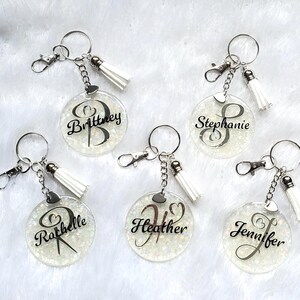 May include: Personalised keyrings with names including "Brittney," "Stephanie," and "Jennifer." Each keyring has a clear, round charm with glitter, a silver chain, a white tassel, and a lobster clasp.