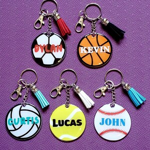 May include: A selection of personalised sports-themed keyrings. Each keyring features a different sport: football, basketball, volleyball, tennis, and baseball. Each keyring has a name printed on it and a tassel.