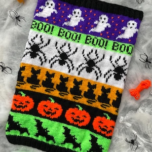 May include: A knitted Halloween-themed blanket with a black border. The design features white ghosts on a purple background, spiders, the word "BOO!" in green, black cats, orange pumpkins, and green bats. Yarn is visible in the corners.