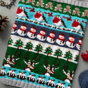Op de afbeelding: Een gebreide kerstcowl met een grijze geribbelde rand. Het ontwerp toont rijen peperkoekenmannen, roodborstjes, sneeuwmannen, kerstbomen, rendieren en kerstpuddingen in rood, wit, groen en blauw.