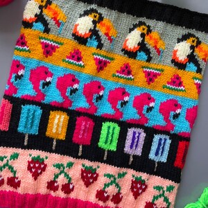 Könnte beinhalten: Ein farbenfrohes Stricktextil mit Mustern von Tukans, Wassermelonenstücken, rosa Flamingos, Eis am Stiel, Erdbeeren und Kirschen. Das Design verwendet verschiedene Farben, darunter Schwarz, Gelb, Rosa, Blau und Grün. Es handelt sich wahrscheinlich um einen Schal oder ein ähnliches Accessoire.
