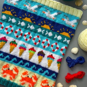 May include: Knitted beach-themed blanket with a yellow border. The design features seagulls, life preservers, ice cream cones, and crabs in shades of blue, white, orange, and yellow. The blanket is made of yarn.