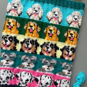 May include: A knitted scarf with a repeating pattern of different dog breeds. The scarf has a teal border and features various dog breeds, including fluffy white dogs, golden retrievers, and dalmatians. The color scheme includes teal, green, pink, and white, creating a vibrant design.