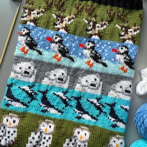 May include: A knitted scarf with a repeating pattern of reindeer, puffins, seals, orcas, and owls. The scarf features bands of green, blue, and grey, with a black border. The animals are in white, black, brown, and orange.