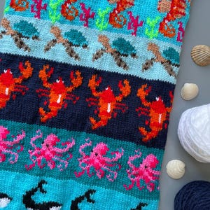 Puede incluir: Textil tejido con un diseño de temática marina. Presenta caballitos de mar, tortugas, langostas, pulpos y orcas en tonos naranja, rosa, azul y turquesa. El diseño se establece sobre un fondo azul marino y turquesa.