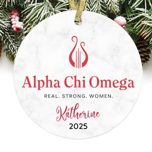 May include: A white, round Christmas ornament with a marble-like texture. The ornament features the text "Alpha Chi Omega" in red, along with a red lyre symbol. Below the text is the name "Katherine" and the year "2025".