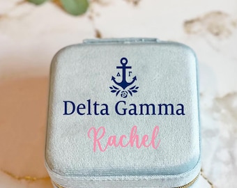 May include: A light blue velvet jewelry box with a gold zipper. The box has a navy blue anchor with a white rose and the letters "ΔΓ" on it. The text "Delta Gamma" and "Rachel" are printed on the box.