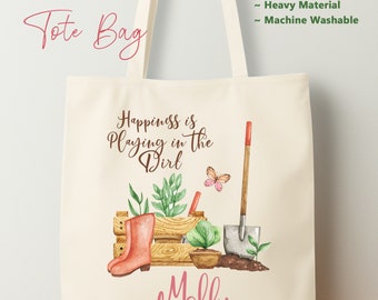 May include: Linen tote bag with a gardening theme. It features a pink gardening boot, a wooden crate with plants, a shovel, and a butterfly. The text on the bag reads "Happiness is Playing in the Dirt" and "Molly".