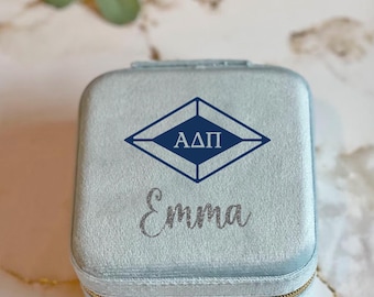 May include: A light blue velvet jewelry box with a gold zipper. The box has a blue diamond shape with the letters "ADPi" in white and the name "Emma" in silver glitter.