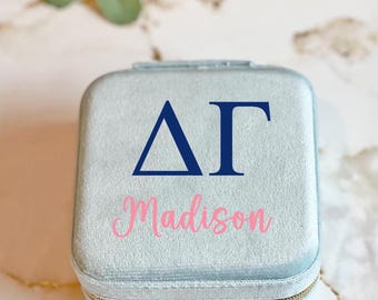 May include: A light blue velvet jewelry box with a zipper closure. The box has a navy blue Greek letter Delta Gamma logo and the name "Madison" written in pink.