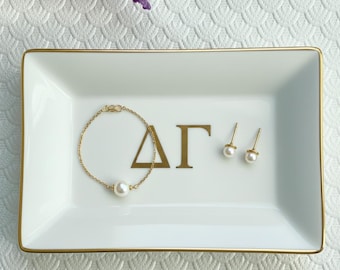 May include: A white rectangular trinket tray with gold trim, featuring a gold bracelet with a pearl, gold earrings with pearls, and gold Greek letters. The tray is on a white textured surface.