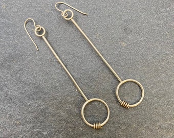 Può includere: Un paio di orecchini dorati con lunghi steli dritti e cerchi circolari alla fine. I cerchi sono collegati agli steli con un piccolo filo attorcigliato.