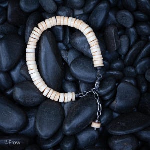 Puede incluir: Una pulsera de color crema compuesta por pequeñas cuentas redondas ensartadas. La pulsera está conectada por una cadena y un cierre de color plateado. El fondo consiste en piedras lisas de color gris oscuro, creando un contraste con el color claro de la pulsera.