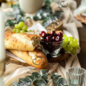 May include: A rustic table setting with a wooden tray, two baguettes, a bowl of cherries, a bunch of green grapes, and two clear glass candle holders. The table is covered in a beige linen tablecloth with eucalyptus greenery draped across it.
