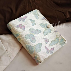 May include: A white fabric journal cover with a repeating butterfly pattern in shades of blue, pink, and green. The cover has a button closure and a textured surface. The journal is rectangular in shape.