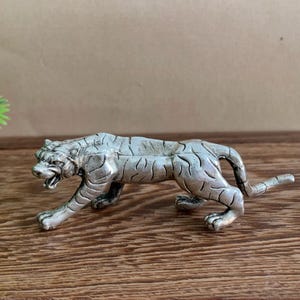 May include: A silver-colored tiger figurine in a walking pose, with its mouth open. The figurine has detailed stripes and is made of a metallic material. The tiger is on a wooden surface, and a green plant is visible in the background.