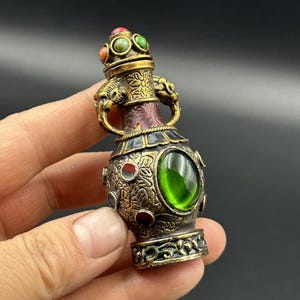 May include: An ornate metal vase in an antique style. The vase has a green gemstone, red and blue accents, and a decorative top with colourful stones. It features two handles with animal head details and intricate patterns.