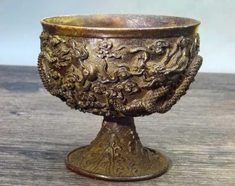 May include: A bronze-colored, ornate chalice with a detailed dragon and cloud design. The cup sits atop a pedestal base with a textured pattern. The chalice has a weathered appearance, suggesting age or antique status.