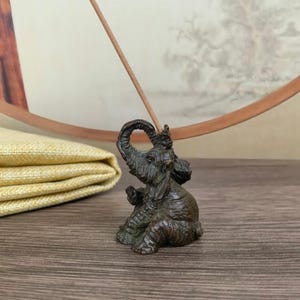 May include: A bronze elephant incense holder with a raised trunk, holding a thin incense stick. The elephant is detailed with wrinkles and sits on a wooden surface. A folded yellow cloth and a decorative circular frame are in the background.