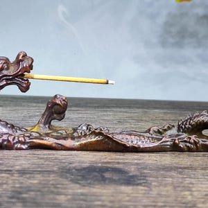 May include: A detailed bronze-coloured dragon incense holder. The dragon's head holds a lit incense stick, with smoke rising. The holder is on a wooden surface, with a blurred background.
