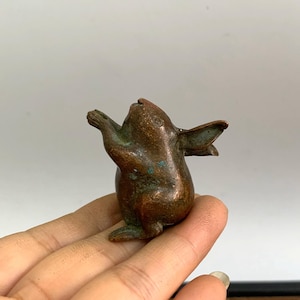 May include: A small, bronze-colored rabbit figurine with outstretched arms and ears. The bunny has a weathered appearance with hints of green patina, suggesting age. The sculpture is detailed and appears to be handmade.