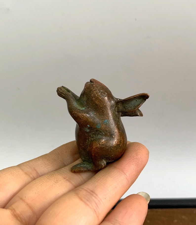 May include: A small, bronze-colored rabbit figurine with outstretched arms and ears. The bunny has a weathered appearance with hints of green patina, suggesting age. The sculpture is detailed and appears to be handmade.