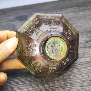 May include: A bronze-coloured, octagonal object with engraved Chinese characters and a central Yin and Yang symbol. The object has a weathered appearance. The background is a wooden surface.