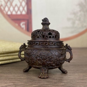 Può includere: Un bruciatore di incenso in bronzo con intricate incisioni a rilievo di draghi e fiori. Il coperchio presenta disegni traforati e un piccolo pinnacolo. Il bruciatore ha due manici a forma di drago e tre piedi. Il design è ornato e dettagliato.