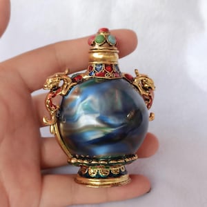 Puede incluir: Pequeña botella decorativa con un marco de metal dorado. La botella tiene un acabado azul, arremolinado y nacarado. Está adornada con joyas rojas, verdes y azules y dos figuras de dragones. La tapa de la botella también está decorada con joyas.
