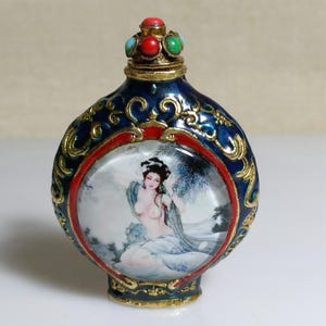 Op de afbeelding: Een sierlijke, blauw en goudkleurige decoratieve fles met een geschilderd beeld van een vrouw. De fles heeft een rode en gouden rand en een juwelen stopper met rode, groene en blauwe stenen. Het ontwerp is ingewikkeld en gedetailleerd.