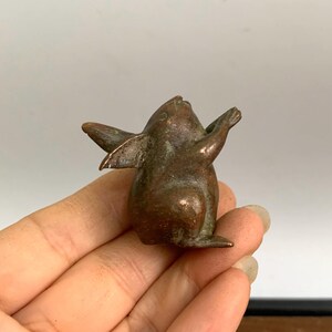 May include: A small, bronze-colored figurine of a creature with wings and outstretched arms. The figurine has a smooth, aged appearance and is held in a person's hand. The creature's wings are slightly spread, and its face is tilted upwards.