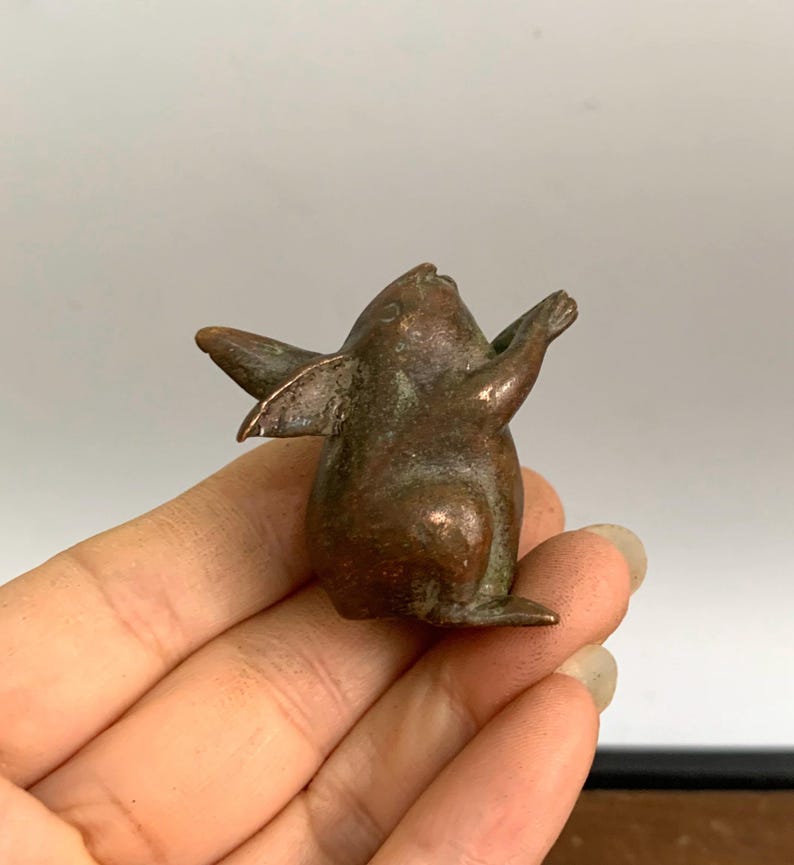 May include: A small, bronze-colored figurine of a creature with wings and outstretched arms. The figurine has a smooth, aged appearance and is held in a person's hand. The creature's wings are slightly spread, and its face is tilted upwards.