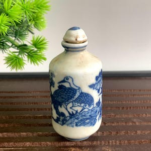 May include: A small, antique white ceramic bottle with a blue bird and floral design. The bottle has a matching blue and white stopper. The bottle is cylindrical with a rounded top and bottom.