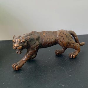 Op de afbeelding: Een klein, bronskleurig metalen tijgerbeeldje in een sluipende houding. Het beeldhouwwerk heeft een gedetailleerd, gestructureerd oppervlak met gegraveerde strepen over het lichaam, een grommende gezichtsuitdrukking met een open bek.
