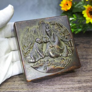 May include: A rectangular, bronze-colored box with a detailed relief carving. The carving depicts two figures in traditional clothing, seated near bamboo and mountains. The box has a slightly worn appearance, suggesting age and use.