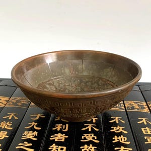 Könnte beinhalten: Eine bronzefarbene Schale mit eingravierten Mustern. Die Schale hat ein breites, flaches Design mit detailliertem Rand und Innenraum. Die Schale wird auf einem Stapel schwarzer Bücher mit goldfarbenen Buchstaben ausgestellt. Der Durchmesser der Schale beträgt ca. 20 cm.