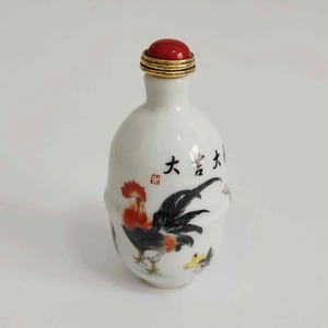 May include: A white ceramic bottle with a red stopper and gold trim. The bottle features a hand-painted rooster with black, red, and yellow details. Chinese characters are written on the bottle.