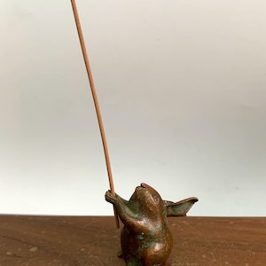 May include: A bronze incense holder shaped like a small animal, holding a brown incense stick. The figurine has a weathered appearance and is set against a neutral background.