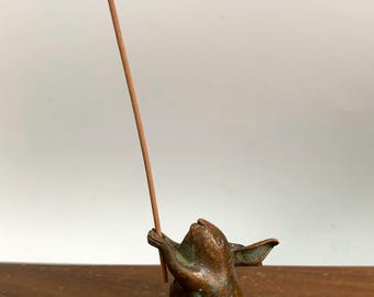 May include: A bronze-colored incense holder in the shape of a small animal, holding a brown incense stick. The figurine has a weathered appearance and is set against a neutral background.
