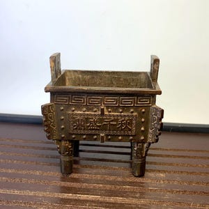 May include: A bronze-colored, rectangular incense burner with a square opening. The burner has decorative patterns and Chinese characters. It features four short legs and two handles. The item is displayed on a wooden surface.